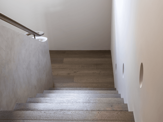 Interior view looking down a modern stairway with broad timber-clad steps, a textured light-grey side wall and a smooth white wall featuring small oval openings. A simple metal handrail follows the stair edge, and the upper landing continues in matching timber flooring. The space is bright, minimal and refined.