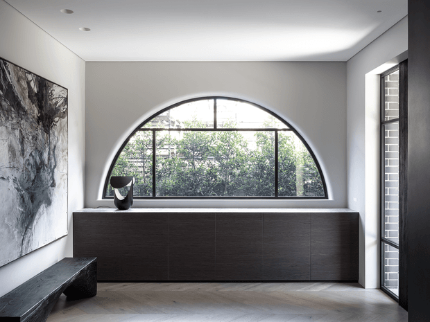 Refined contemporary interior featuring a large arched steel-framed window, dark timber built-in cabinetry, pale herringbone timber flooring, white plaster walls, and a minimalist sculptural bench and artwork. Natural greenery is visible outside the window.