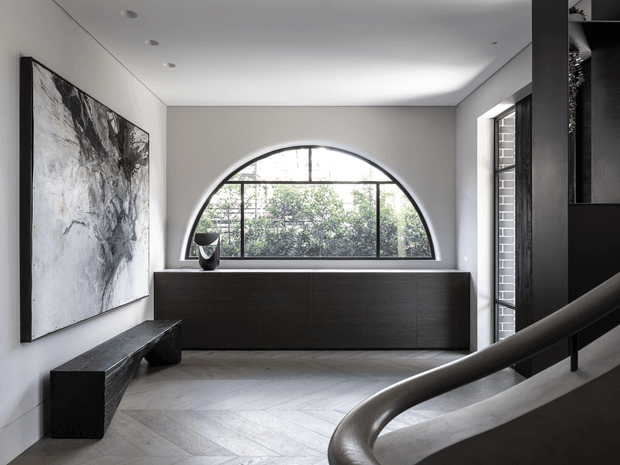 A refined contemporary entry hall with a large arched steel-framed window, dark built-in storage cabinet, sculptural dark bench, abstract wall artwork, pale herringbone flooring, and a curved stair handrail in the foreground. The space has a restrained monochromatic palette and abundant natural light.