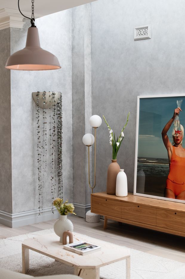 A refined living room with textured grey plaster walls, decorative white ceiling mouldings and skirting, a suspended terracotta pendant light, timber mid-century-style media console, sculptural floor lamp, ceramic vessels, and a large framed artwork. A trailing indoor plant adds greenery beside the television, while a pale stone coffee table and light rug complete the softly styled space.