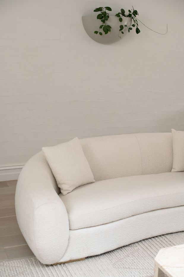 A softly styled living room corner featuring a large rounded boucle sofa in warm off-white, matching cushions, a pale textured rug, light timber flooring, and a white-painted brick wall. A simple wall-mounted planter with trailing greenery adds a natural accent.