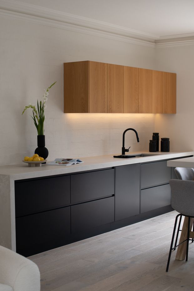 A refined contemporary kitchen featuring matte black lower cabinetry, warm natural timber overhead cabinets, a pale stone benchtop, black gooseneck tapware, and subtle under-cabinet lighting. The room has white painted brick walls, ornate ceiling cornices, pale timber flooring, and a partially visible upholstered bar stool. Minimal styling includes black vessels, greenery, fruit, and reading material.