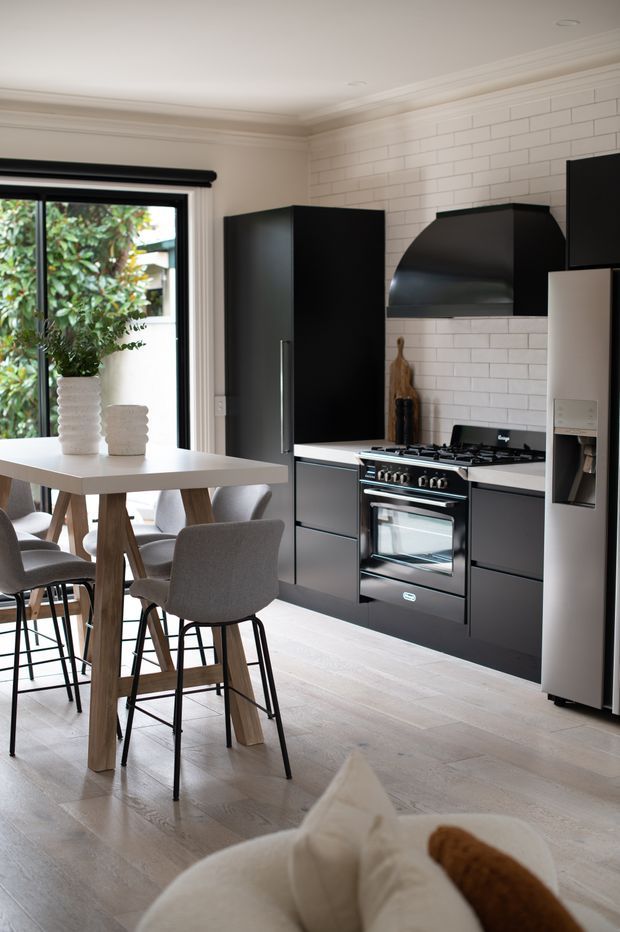 A bright contemporary kitchen-dining space with matte black cabinetry, a black freestanding range and sculptural rangehood set against a white subway-tile backsplash. A pale timber dining table with upholstered bar stools sits beside large black-framed glazing, while light timber flooring, restrained cornices and neutral furnishings create a refined, airy interior.