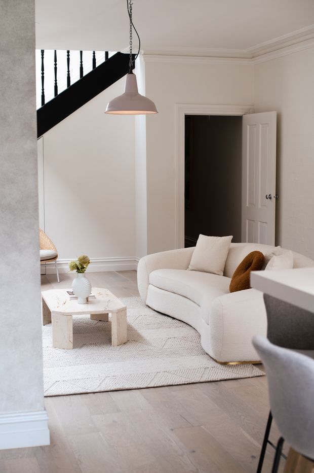 A bright, refined living room with white walls and traditional decorative mouldings, pale timber flooring, and a curved cream sofa arranged around a light stone coffee table. A textured neutral rug, sculptural pendant light, and black-painted staircase balustrade add contrast. A doorway and partial dining or seating area are visible beyond the room.