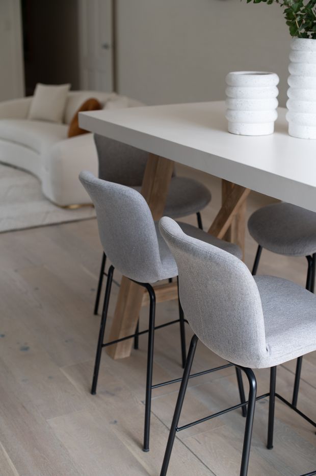 A softly styled open-plan dining and living area. A white rectangular island-height dining table with a thick top and timber angled legs is surrounded by grey upholstered stools with black metal frames. Pale timber flooring, a white curved sofa, neutral walls, and white textured vases with greenery create a calm, contemporary interior.