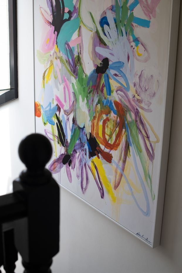 Close-up interior view of a large, colourful abstract floral artwork mounted on a light wall beside a dark timber stair balustrade. The painting features energetic brushstrokes in pink, turquoise, blue, green, purple, yellow, orange and black, with natural light entering from the left.
