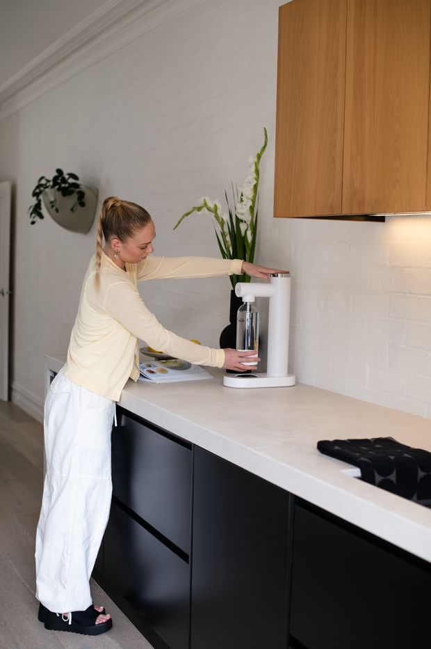 A contemporary kitchen interior with a long pale stone benchtop, matte black base cabinetry, warm timber overhead cupboards, and a white painted brick backsplash. A person is using a compact white countertop appliance on the island, with understated decor and indoor plants. The space has a clean, refined, softly warm aesthetic.