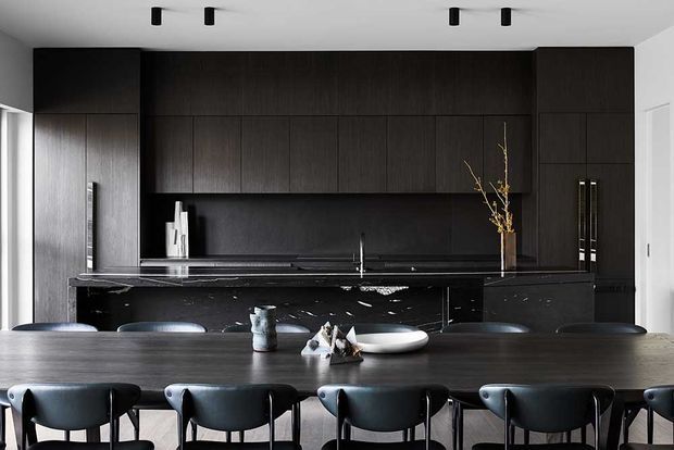 A sophisticated open-plan kitchen and dining area with dark floor-to-ceiling timber cabinetry, a black stone island, integrated appliances, and a long dark timber dining table with black upholstered chairs. Minimal black track lighting and a restrained monochromatic palette create a refined contemporary atmosphere.