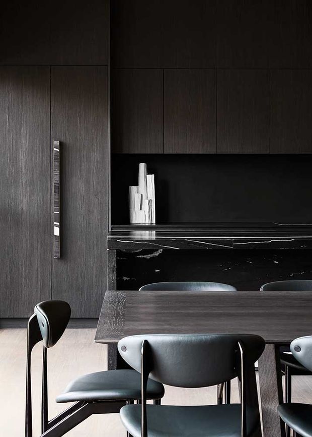 Dark, sophisticated open-plan kitchen and dining area with charcoal timber-look cabinetry, a black stone island or benchtop, and a dark wood dining table surrounded by sculptural black leather chairs. A reflective black splashback and minimal white decorative objects add contrast, while pale flooring balances the moody palette.