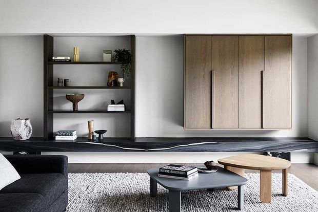 A refined contemporary living room featuring a dark built-in entertainment and display wall, a large floating timber storage cabinet, dark stone-like shelving and bench surfaces, and minimalist coffee and side tables. A black sofa and textured light rug create a balanced monochrome and warm-wood palette.