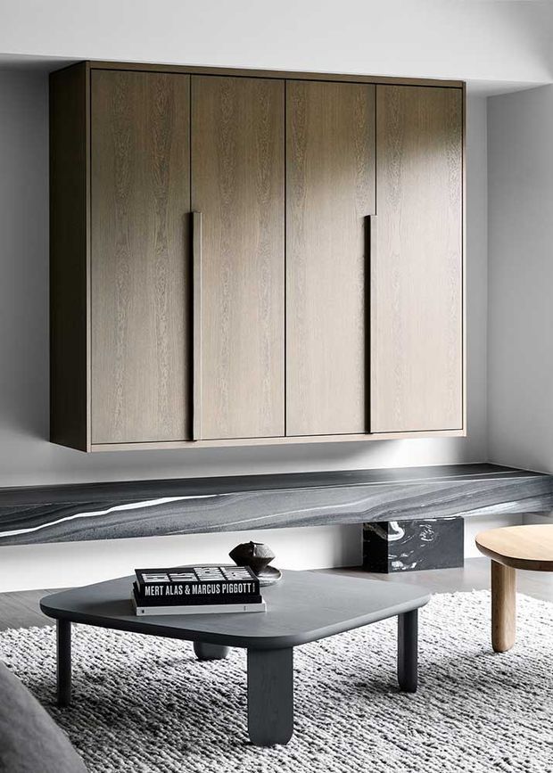 Minimalist contemporary interior featuring a wall-mounted, light timber built-in cabinet with tall flush doors and integrated vertical pulls. Below it is a long dark veined stone ledge or benchtop, with a low dark coffee table, textured light rug, and a small round timber side table. The room has white walls, restrained detailing, and a monochromatic palette with natural wood accents.