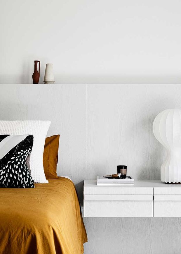 A styled bedroom vignette featuring a mustard-gold bedspread, layered white and black patterned pillows, and a floating white timber-textured bedside cabinet. The room has white textured wall panelling, a simple ledge displaying ceramic vessels, and a sculptural white table lamp, creating a clean, high-contrast contemporary look.