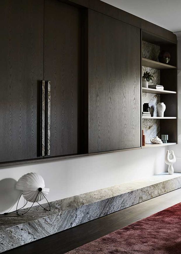 Close-up of a sophisticated built-in living room media and storage wall. Dark timber cabinetry with long vertical handles sits above a long pale natural-stone console, with open display shelving, books and sculptural decor. A dark timber floor and burgundy rug add contrast, creating a moody, refined interior.