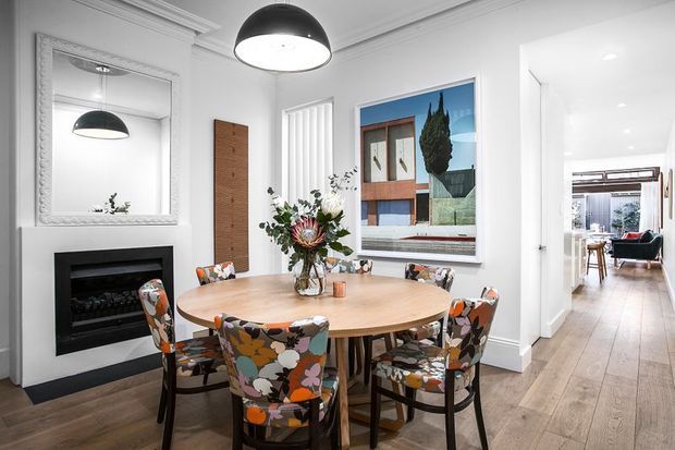 Bright, refined dining room with a large round timber dining table surrounded by patterned upholstered chairs. The room features white walls with decorative ceiling cornice, a black pendant light, ornate wall mirror, built-in black fireplace, timber flooring, and a large framed artwork. An open doorway leads to a bright adjoining living area.