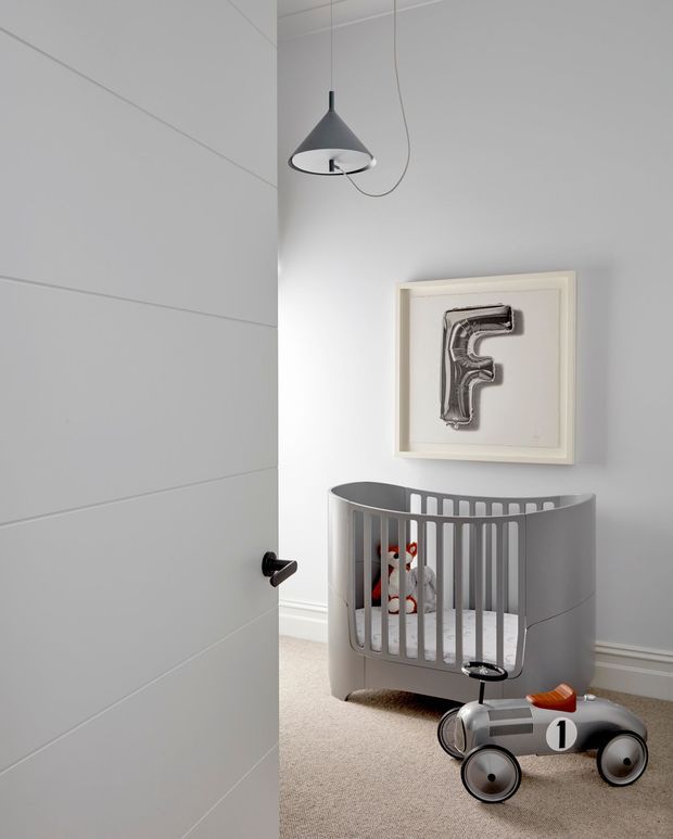 A softly lit contemporary nursery viewed through a partially open white door. The room features a grey modern cot, a silver-and-black ride-on toy car, a minimalist pendant light, pale grey walls with horizontal panel lines, beige carpet, and framed monochrome artwork.