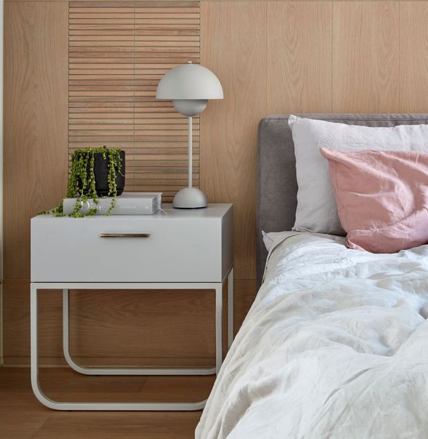 A close-up of a contemporary bedroom showing a white bedside table with rounded metal legs, a sculptural white dome table lamp, books, and a trailing plant beside a partially visible upholstered bed. The room features warm light timber wall panelling and flooring, including a slatted timber feature panel, with white bedding and soft pink and grey cushions.