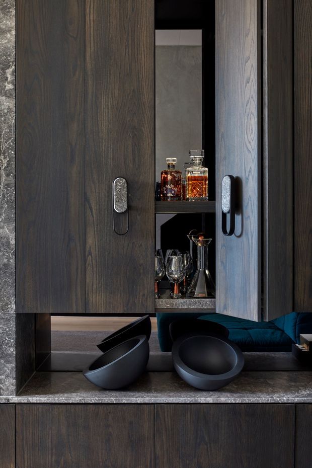 Close-up of a sophisticated built-in drinks cabinet or home bar, featuring dark timber veneer cabinetry, integrated vertical handles, open shelving with decanters and glassware, a dark stone benchtop, and a mirror-backed niche. Decorative black bowls sit on the counter, with a dark teal upholstered seating area reflected in the mirror. The composition is moody, refined, and highly detailed.