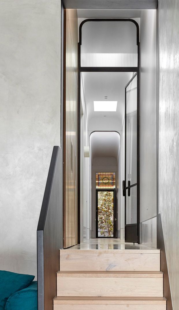 A narrow, refined interior stairway leading into a long hallway. Pale timber stairs and flooring contrast with textured light walls, dark metal handrails and trim, glazed internal doors, and a series of arched openings. A stained-glass panel and garden view appear at the far end, while a teal upholstered surface is partially visible at the lower left.