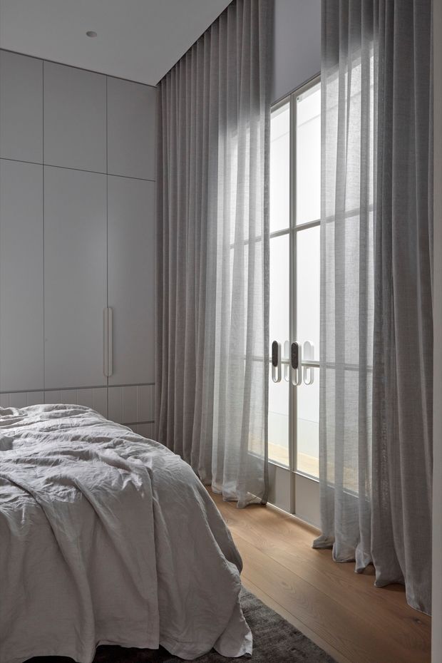 A calm, minimalist bedroom with a neatly built-in wall of pale cabinetry, a partially visible bed dressed in light grey bedding, and full-height sheer grey curtains over tall glazed doors or windows. Warm timber flooring and soft natural light create a quiet, refined atmosphere.