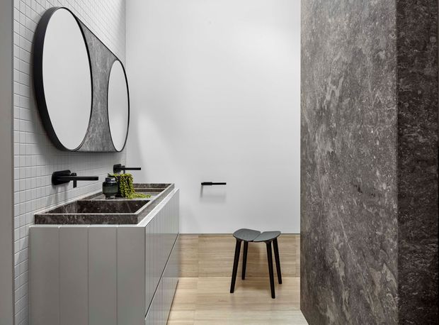 Contemporary bathroom with a long floating double vanity, twin round mirrors, black wall-mounted fixtures, pale tiled walls, light timber flooring, and a dramatic dark stone feature wall. A small black stool and minimal greenery add restrained detail.