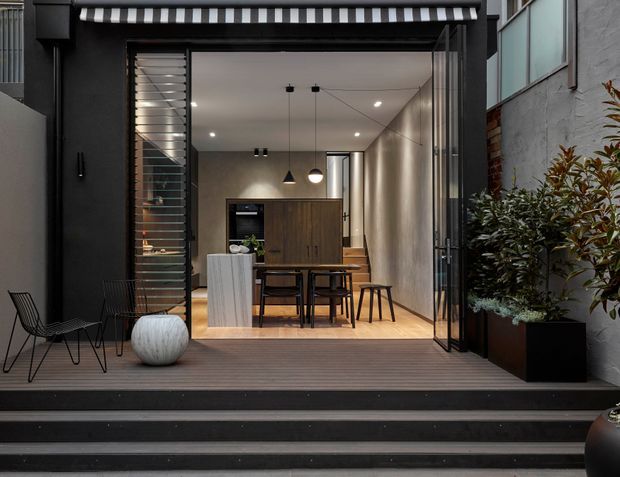 A compact contemporary urban dining area viewed from an outdoor terrace through fully opened glazed folding doors. The interior features a timber dining table and chairs, a dark built-in kitchen or storage wall, pendant lighting, recessed ceiling lights, and warm timber flooring. Dark exterior cladding, a striped awning, black decking and steps, metal-framed outdoor chairs, and large potted planting frame the opening.