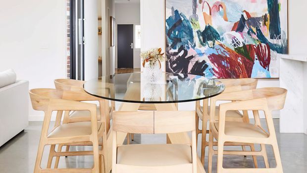 Bright contemporary dining area with a large round glass-topped dining table, light timber dining chairs, and a pale timber central pedestal. A colorful abstract artwork forms a feature wall, while a white sofa and open hallway are visible in the background. The room has a clean, airy, neutral fit-out with warm wood accents.