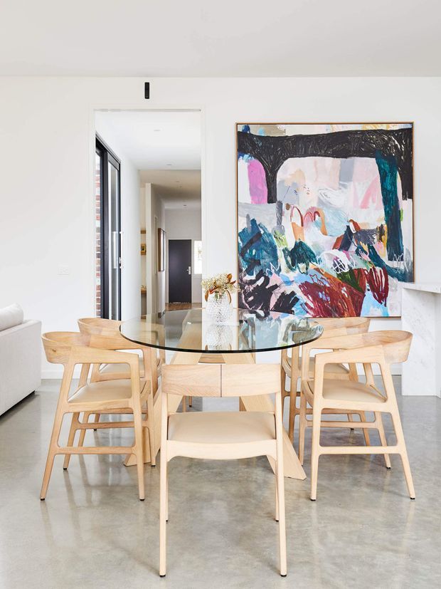 Bright contemporary dining area with a round glass dining table surrounded by light timber chairs. The room features polished concrete flooring, white walls, a large colourful abstract artwork, extensive glazing and a minimalist open-plan connection to the hallway and living area.