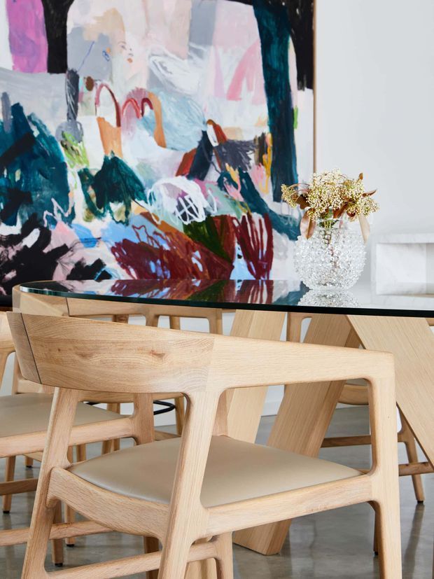 A contemporary dining area with a large abstract multicolour artwork behind a rectangular glass-top dining table. The table is surrounded by sculptural light timber dining chairs with tan upholstered seats. A clear textured glass vase with delicate dried flowers sits on the table, creating a refined yet expressive interior.