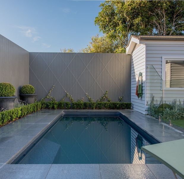 A compact landscaped courtyard with a rectangular swimming pool beside a light weatherboard-clad house. The pool is surrounded by dark grey tiled or stone paving, clipped hedges, potted topiary, and a tall grey privacy fence with a geometric diagonal pattern. The scene has bright natural daylight and a clean contemporary-residential feel.