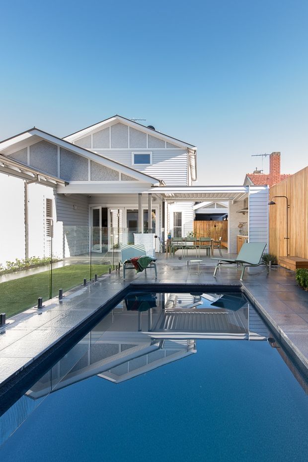 Bright exterior view of a contemporary residential courtyard with a long rectangular swimming pool, paved pool deck, glass pool fencing, outdoor lounge furniture, a covered entertaining area, and light weatherboard-clad houses surrounding the space. The composition highlights the pool and private outdoor living area, with pitched gable roofs and timber privacy fencing visible in the background.