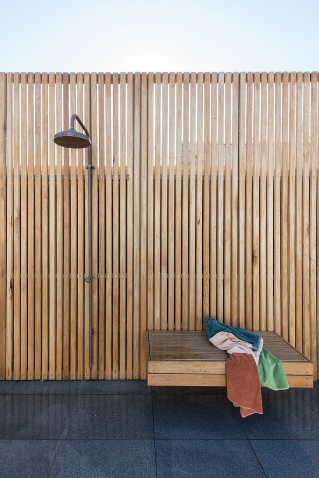 A minimalist outdoor shower area with a vertical timber slat privacy screen, dark metal shower fixture, built-in timber bench, folded towels, and large dark floor tiles. The warm wood contrasts with the charcoal flooring and pale sky.
