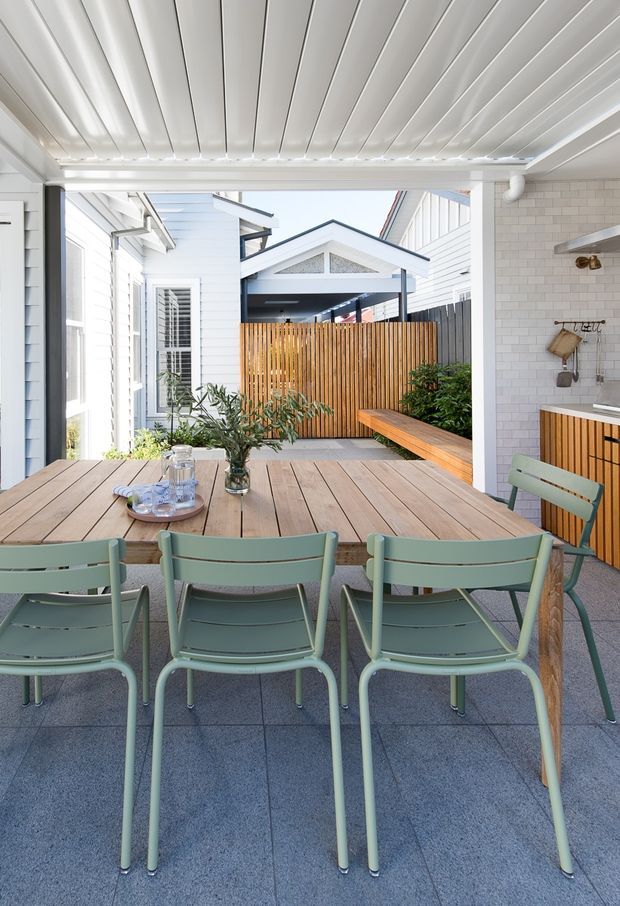 A bright covered outdoor dining courtyard with a large timber dining table and pale green chairs. White weatherboard walls, a white slatted ceiling, tiled flooring, a white tiled outdoor kitchen with timber cabinetry, and a timber privacy screen surround the space. The courtyard opens toward a small gabled structure and landscaped planting.