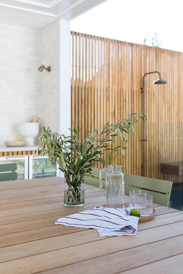 Bright, airy interior showing a timber dining table in the foreground with green dining chairs, a glass vase of leafy branches, a water carafe and glasses. Behind it is a compact kitchen or entertaining area with white tiled walls, a dark curved faucet, pale cabinetry, and a full-height vertical timber slat screen. A roof skylight or clerestory-like opening brings in abundant natural light.