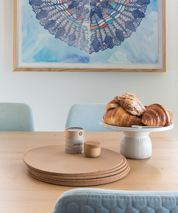 A softly styled dining area with a light timber table, pale blue upholstered dining chairs, and a sculptural white ribbed pedestal bowl filled with pastries. A round layered timber serving board holds a takeaway coffee cup and small wooden vessel, while a large framed blue-toned abstract artwork hangs on the pale wall behind.