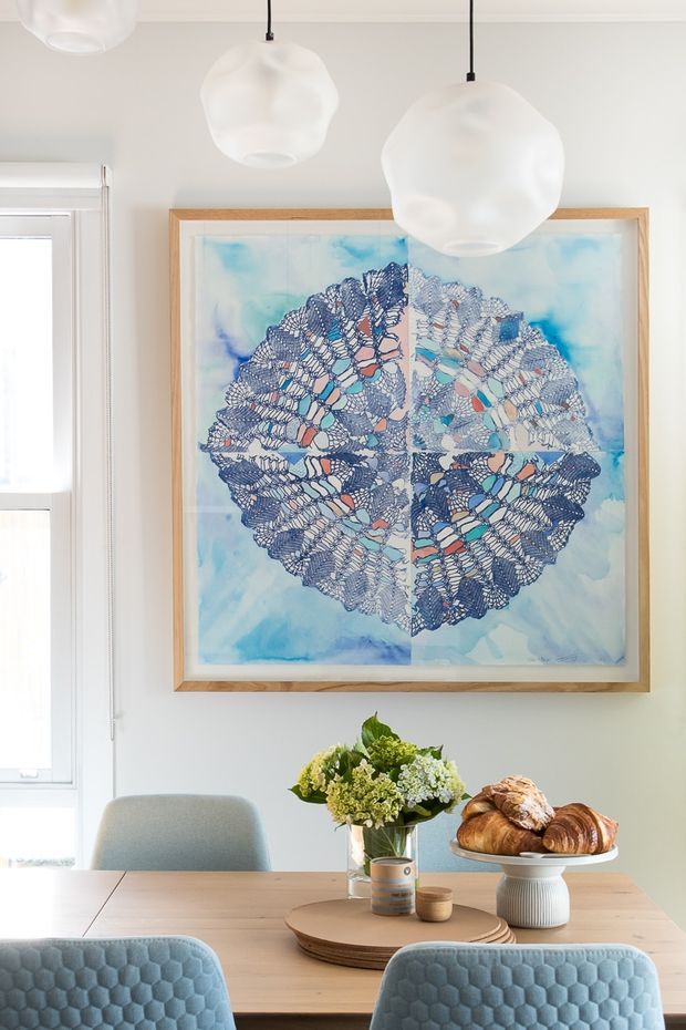 Bright, airy dining area with a light timber dining table, upholstered blue-grey chairs, a large framed blue abstract artwork, and sculptural white pendant lights. A floral arrangement, plates, and pastries create an inviting breakfast setting beside a tall window.