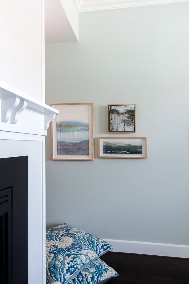 A softly lit bedroom corner with pale blue-grey walls, white decorative crown moulding and a built-in white fireplace surround with a black firebox. Three framed landscape artworks hang above the dark timber floor, while blue patterned bedding is visible at the lower edge.