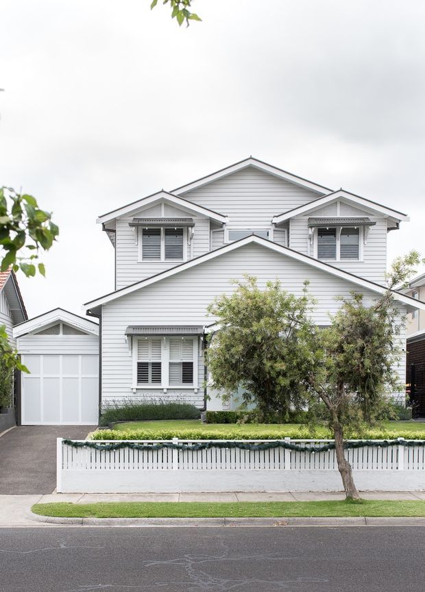 Front exterior view of a two-storey suburban home with a layered gabled roofline, white horizontal weatherboard cladding, decorative gable detailing, grey-trimmed windows with shutters, an attached white garage, and a low white picket-style boundary fence. A small landscaped front garden and street frontage are visible.
