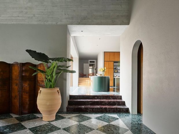 A refined residential entry hall with a dramatic geometric checkerboard stone floor, dark veined marble steps, textured plaster walls, and a sculptural arched opening. A large potted tropical plant and antique wooden screen add warmth, while the view beyond reveals a contemporary kitchen or living area with warm timber cabinetry and a teal island.