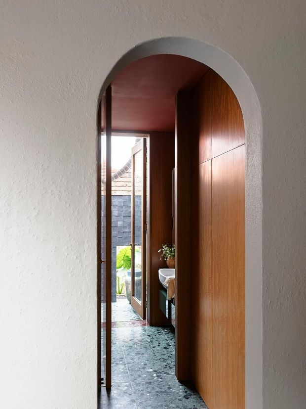 A narrow interior passage framed by a rounded archway, with warm timber wall panelling and a matching timber door on the right. Dark terrazzo flooring leads toward a glazed exterior door and a small courtyard beyond. The space has white textured plaster walls and ceiling, warm natural light, and a refined, minimalist character.
