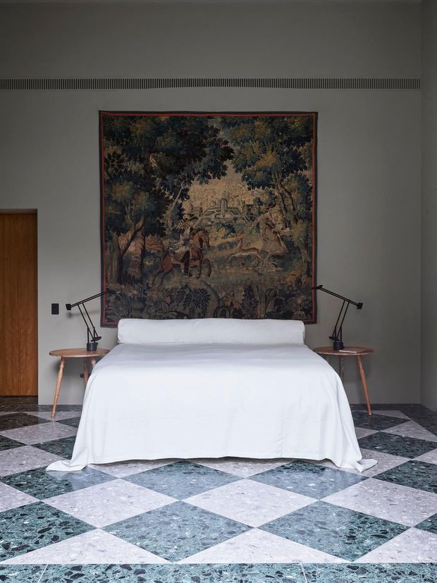 A refined bedroom with a centrally positioned bed dressed in white linen, framed by slim timber bedside tables and articulated black task lamps. A large vintage-style tapestry forms the feature wall above the bed, while the room is grounded by a bold checkerboard floor of pale and dark green terrazzo or stone tiles. The minimalist grey walls and concealed linear ventilation grille create a clean, architectural backdrop.