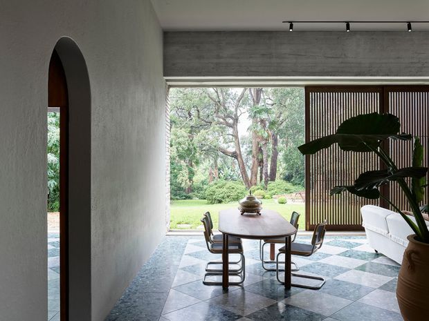 A refined dining area with a dark patterned stone-tile floor, textured light plaster walls, and a sculptural arched opening. A compact oval dining table with cantilevered chairs sits beside expansive glazing overlooking a lush garden. Vertical timber screening, a white sofa, indoor plants, and discreet track lighting create a contemporary indoor-outdoor composition.