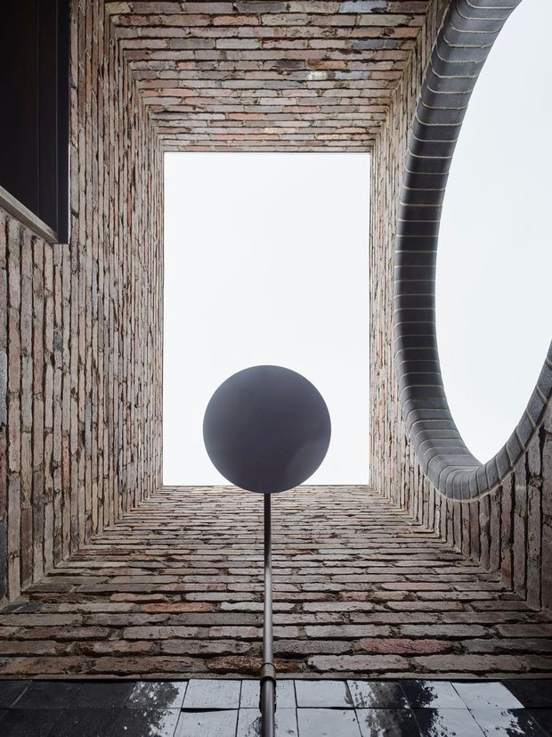 Upward-looking view within a tall, narrow brick-lined architectural void or courtyard. A rectangular opening to the overcast sky is framed by layered exposed brickwork, with a large curved dark metal duct or pipe entering from the upper right. A spherical dark pendant or sculptural light hangs centrally above a reflective dark tiled floor.