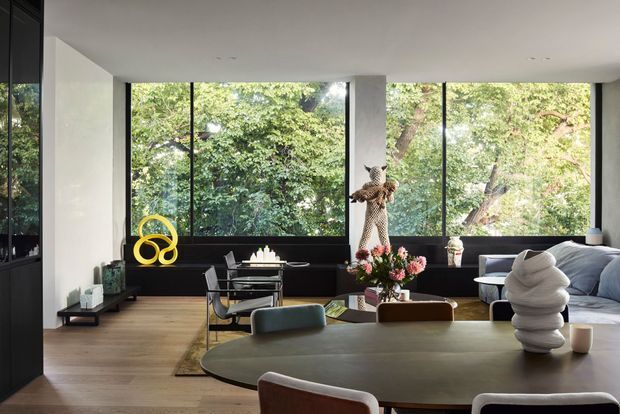A sophisticated contemporary open-plan living and dining room with large floor-to-ceiling windows overlooking dense greenery. The room features a dark built-in window bench and cabinetry, light timber flooring, a large rounded dining table, sculptural decor, a modern sofa, and refined modern furnishings.