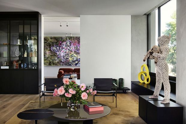A sophisticated contemporary living room with warm timber flooring, black built-in cabinetry and display shelving, a low glass coffee table, and mid-century-style seating. A large abstract floral artwork, sculptural artwork, and potted flowers add colour against a light neutral wall palette. Full-height glazing brings in abundant natural light and views of greenery.
