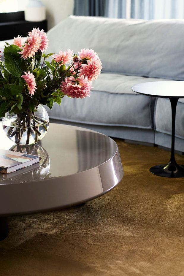 A softly lit contemporary living room featuring a pale grey sofa, a large glossy sculptural coffee table, a slim black side table, and a glass vase of pink flowers. Brown carpet and cool blue-grey furnishings create a refined, tonal interior.