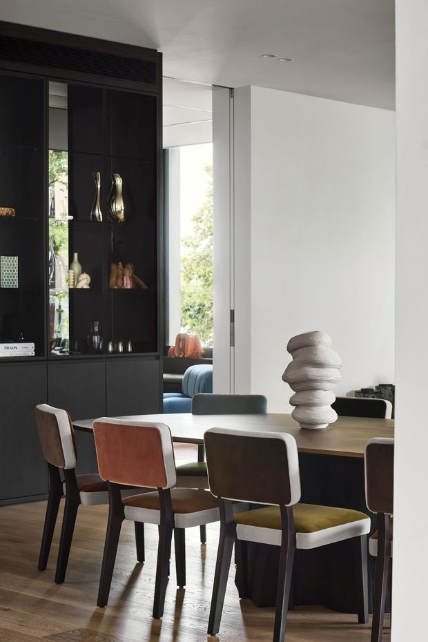 A refined contemporary dining room with a long timber dining table and upholstered dining chairs in contrasting neutral and muted tones. A dark built-in display and storage wall occupies the left side, while a large opening provides views toward a bright adjacent sitting area and greenery outside. The room has white walls and ceiling, timber flooring, recessed downlights, and sculptural decorative objects.