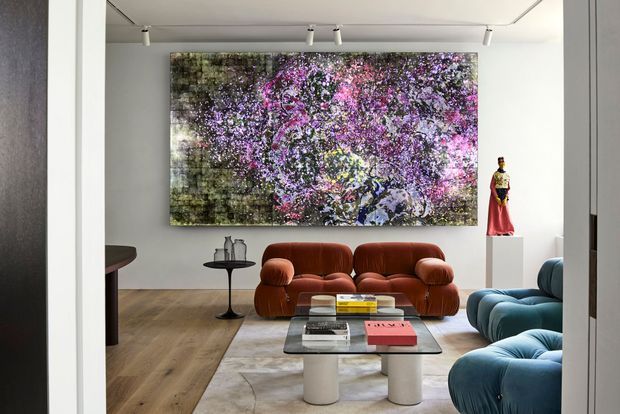 A refined contemporary living room viewed through a doorway, featuring a large abstract floral artwork, sculptural rust-coloured sofa, blue curved lounge seating, a glass coffee table, light timber flooring, and minimalist display pieces. Track lighting and clean white walls create a gallery-like setting.