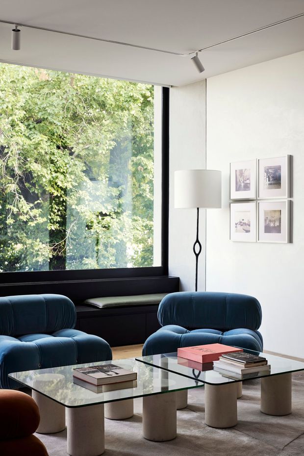 A refined contemporary living room with a large black-framed picture window overlooking dense greenery. Two deep blue upholstered lounge chairs face a low glass coffee table supported by cylindrical stone or concrete legs, with books arranged on top. A black built-in window seat, sculptural floor lamp, track lighting, and four framed artworks complete the calm, gallery-like interior.