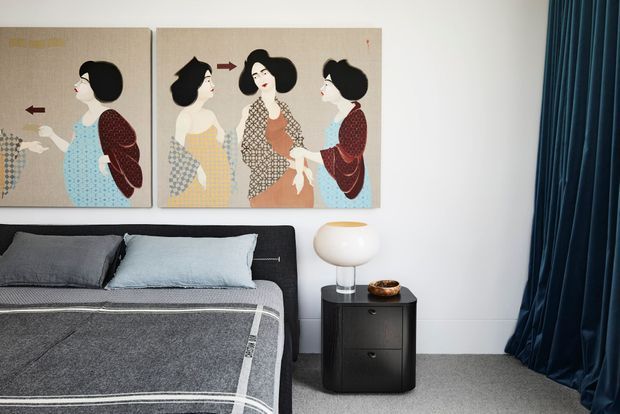 A refined bedroom vignette featuring a low dark upholstered bed with layered grey and pale blue bedding, a compact black bedside table, and a sculptural white table lamp. Two framed vintage-style fashion artworks hang above the bed, while full-height deep blue curtains frame the right side. The palette is muted and elegant with a contemporary, slightly retro character.