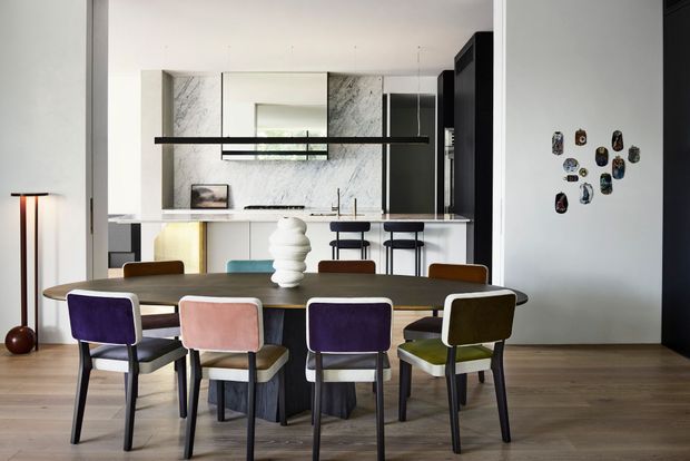 A contemporary open-plan dining area with a large oval dining table and colourful upholstered chairs, positioned in front of a sleek kitchen. The kitchen features dark cabinetry, a veined stone backsplash, a pale island or peninsula, and a linear black pendant light. Light timber flooring, white walls, and sculptural wall decor create a refined, modern interior.